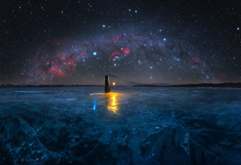 Flat, dark blue ice with a figure and a yellow light with arching Milky Way overhead.