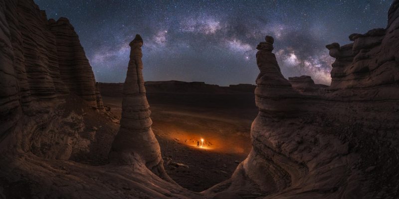 Two conical stone pillars and a bowl of rock with a person standing inside and Milky Way overhead.