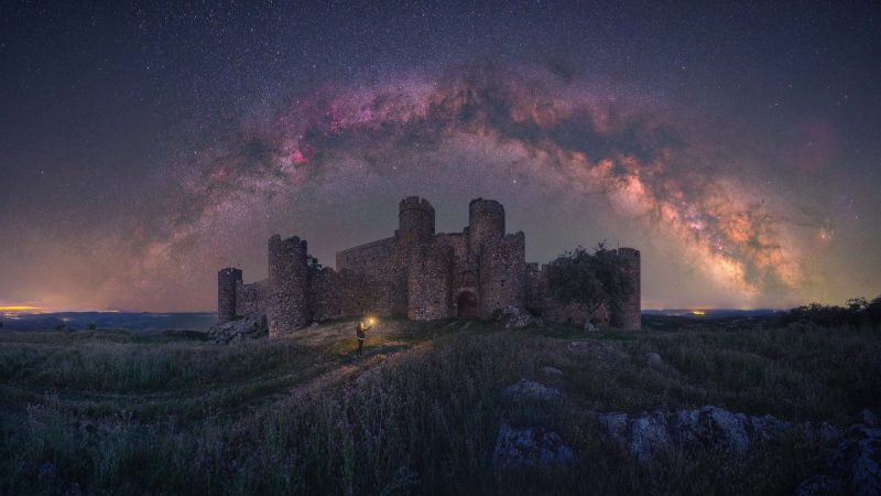 Large stone castle with many towers and with Milky Way arching overhead.