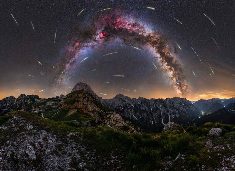 Numerous thin streaks of light in the sky and arching Milky Way over mountainous terrain.