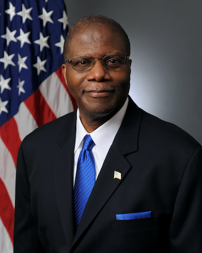 Middle-aged man in suit and tie with American flag behind him.