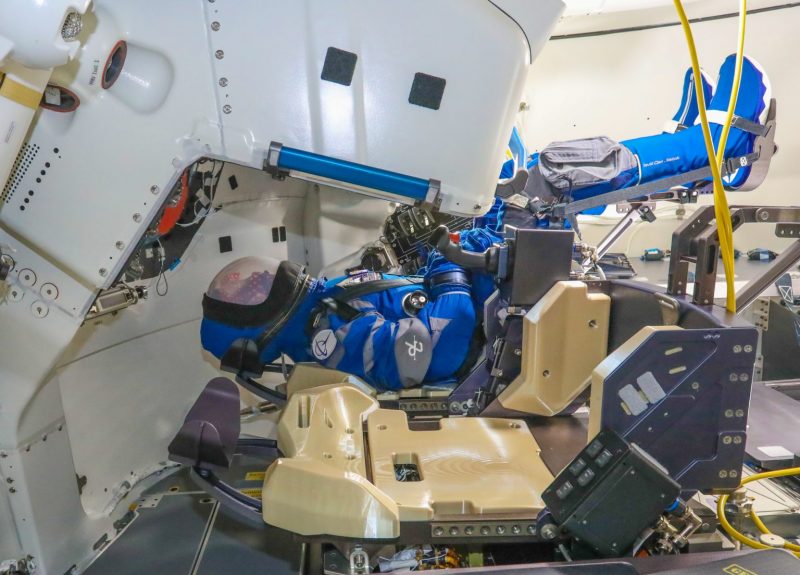 A mannequin (dummy) lying in the pilot's seat in the complex interior of a space capsule.