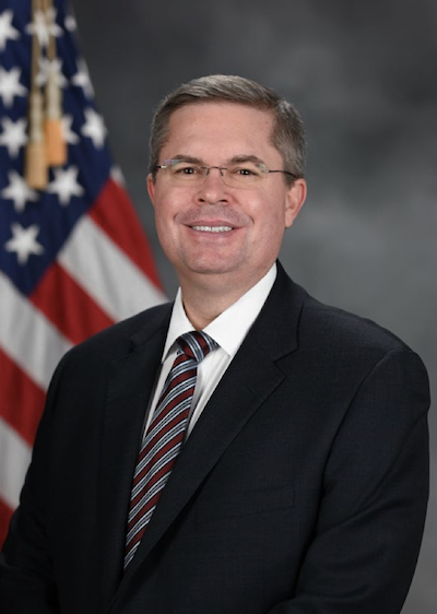 Smiling middle-aged man in suit and tie with American flag behind him.