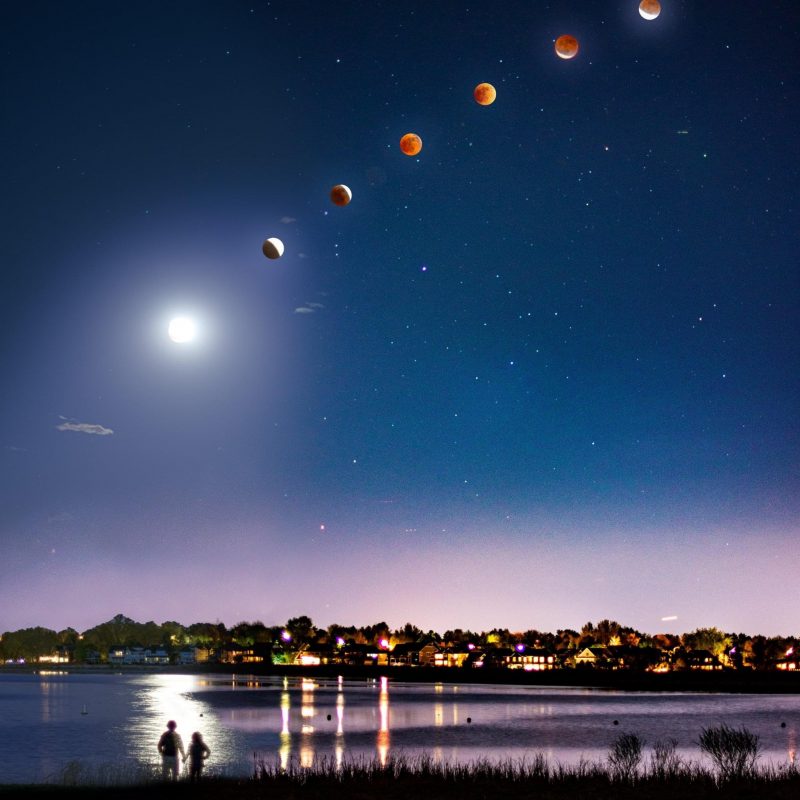 Stages of lunar eclipse over city and pond, with 2 people looking at eclipse.