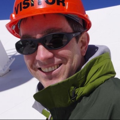 Smiling man with sunglasses and helmet, with snow behind him.