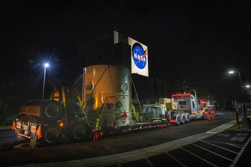 Large round container on long truck platform, with NASA logo on top, at night.