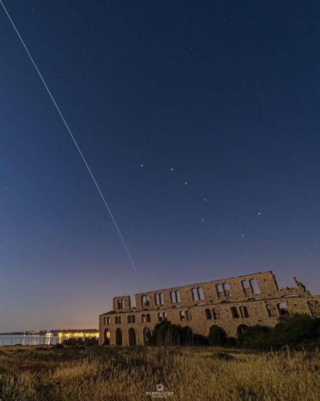 White line in the deep blue sky, along with Big Dipper, with ruins of a stone building below.