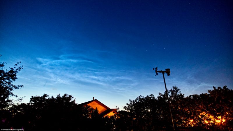 Darkening blue sky with white ripply clouds, below them, there is a house with a weathervane on top and some trees.