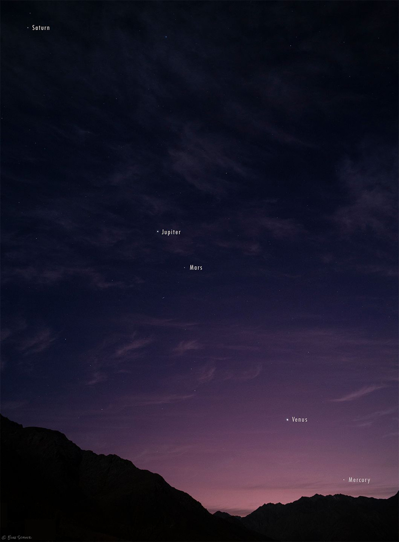 Dark sky with 5 planets lined up in the sky with dark mountains in the foreground.