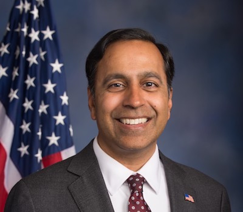 Smiling man in suit and tie with American flag behind him.