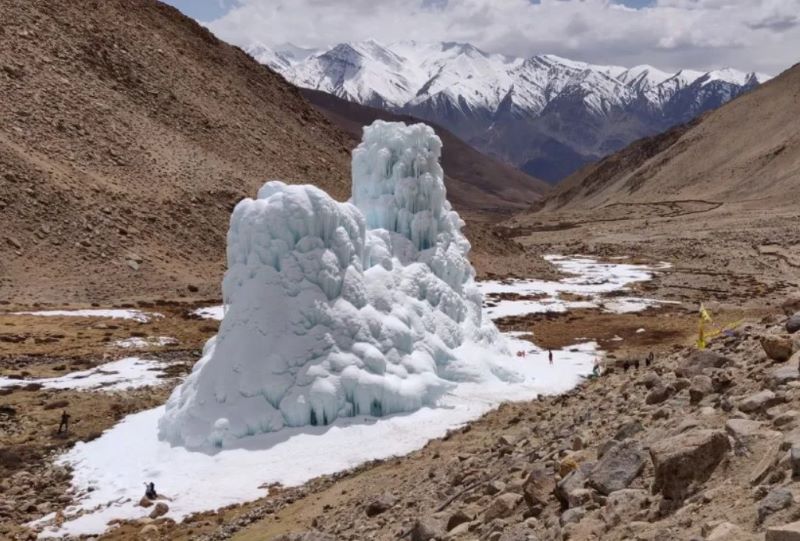 Large white cone-shaped mound of ice in barren valley among mountains, tiny people at base.