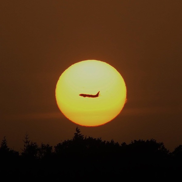 Large, bright yellow circle of sun in deep orange sky with jet plane crossing in front of it.