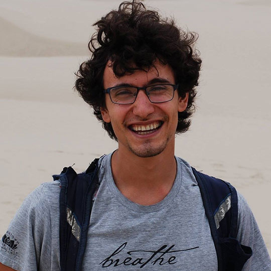 Smiling young man with lots of curly black hair, wearing a backpack in the desert.