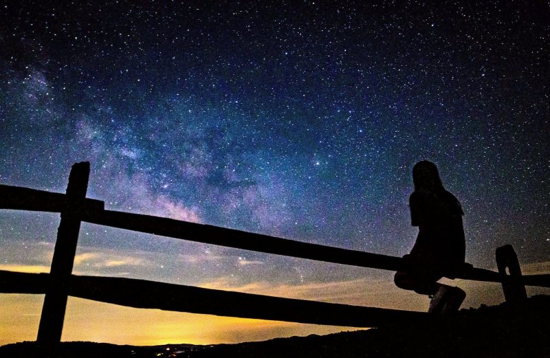 Kid sitting on a rail fence and looking at the Milky Way.