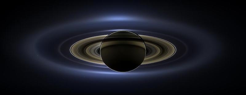 Dark-looking planet surrounded by brilliant rings and a larger, outermost translucent bluish ring.