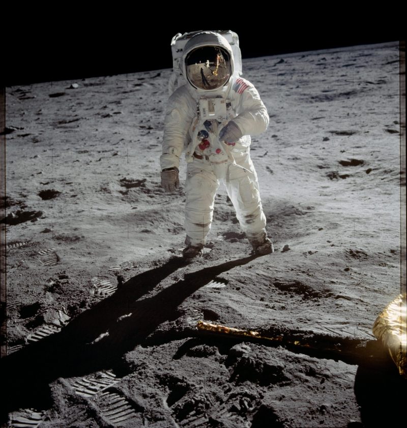 Astronaut in white space suit, helmet and backpack stands on gray, rocky surface of moon.