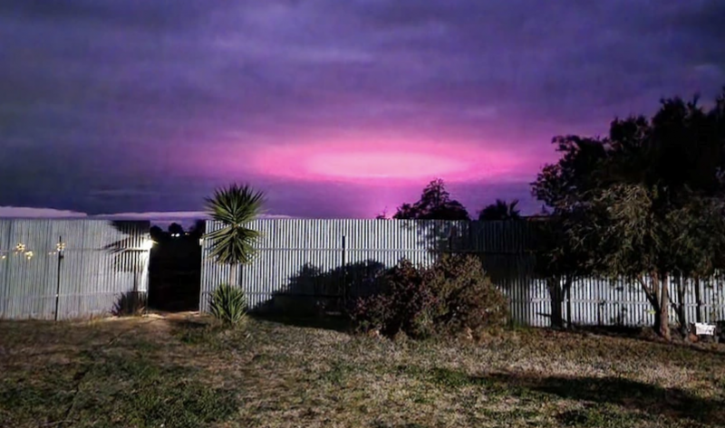 Pink oval in a purple sky behind a white corrugated metal building.