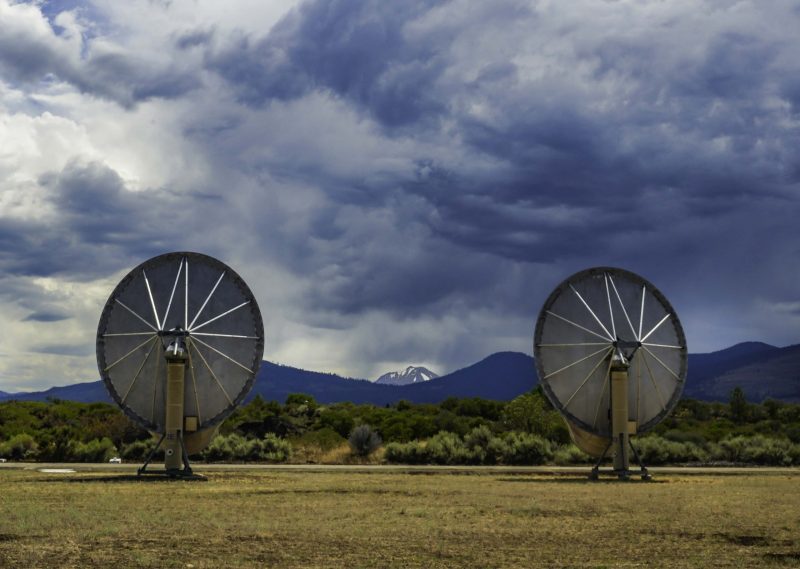 Cloudy sky and mountains in the background with 2 radio dishes in the foreground.