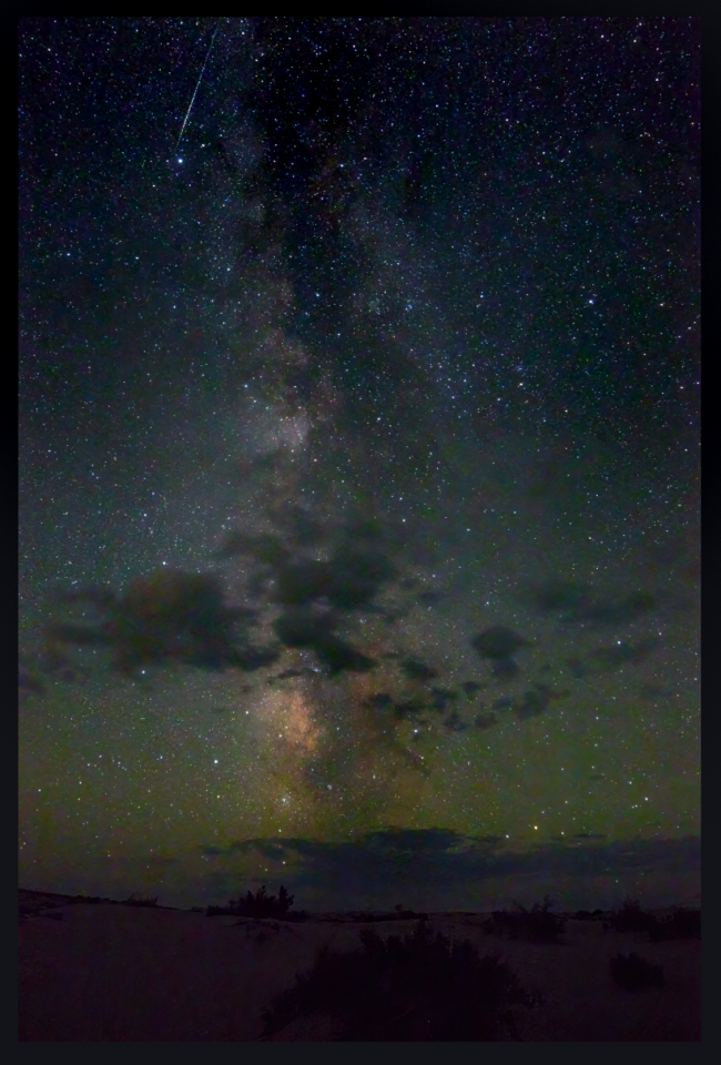 Vertical Milky Way with some foreground clouds, and high, thin streak of meteor.