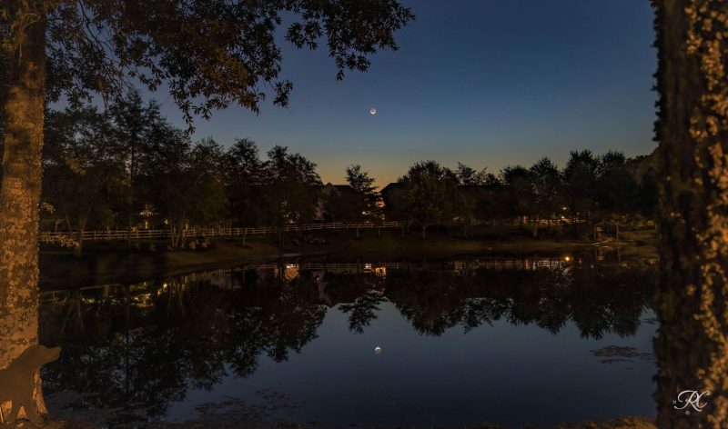 Crepúsculo matutino con luna creciente y Venus con árboles que rodean un lago.