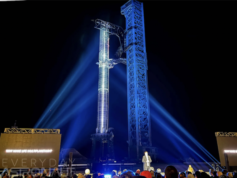 A very tall shiny cylindrical object stands upright with blue lighting casts upon it. A man stands below on a stage while a crowd of people faces him.