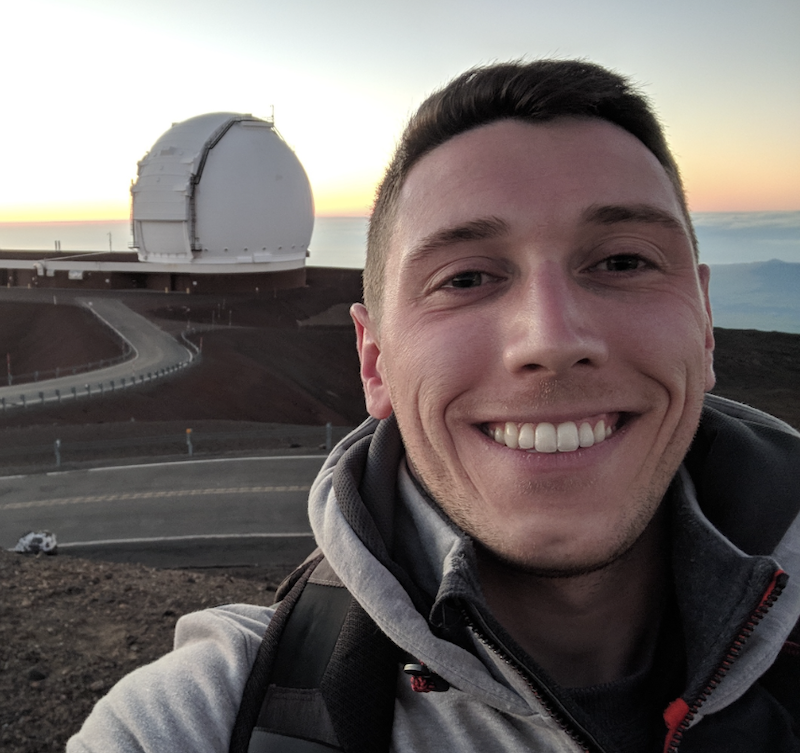 Smiling man with short hair and telescope observatory and roads behind him.