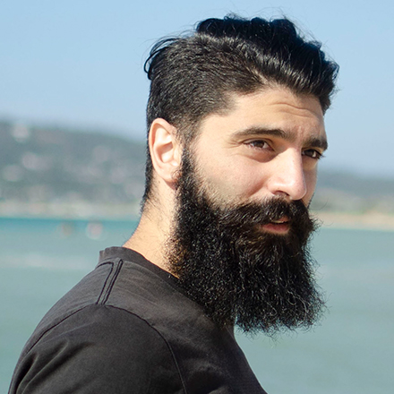 Man with dark hair and large dark beard looks over his shoulder, with sea in background.