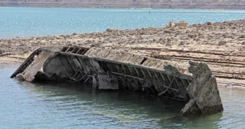 Large half-sunken boat lying on its side in shallow lake water by barren rocky shoreline.