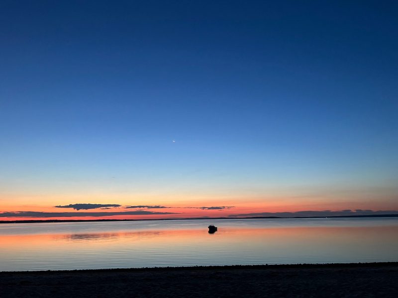 Sunrise colors reflected on water with crescent moon above.