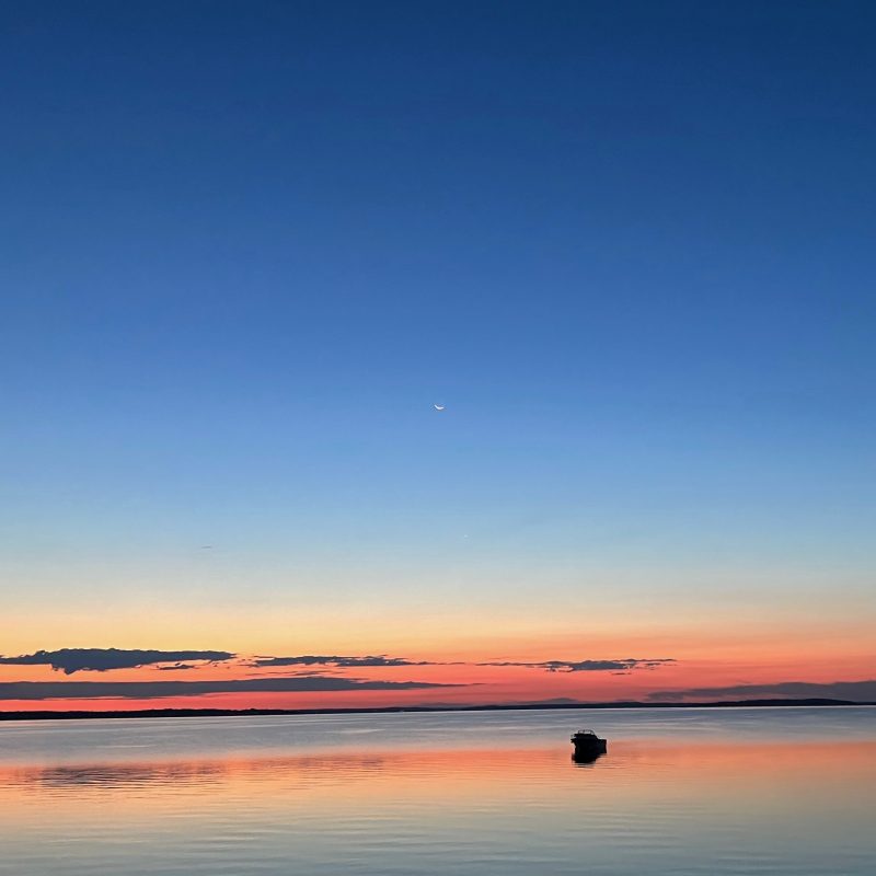 Sunrise colors reflected on water with crescent moon above.