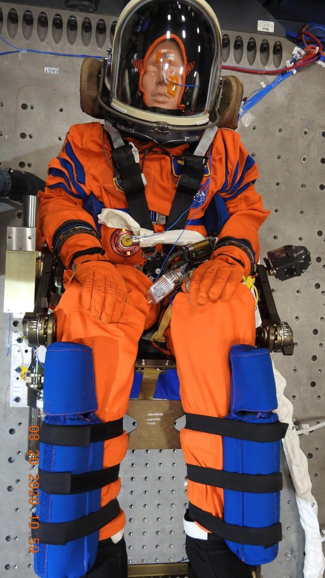 A seated orange-suited mannequin with space helmet on, with a belt and wires around.