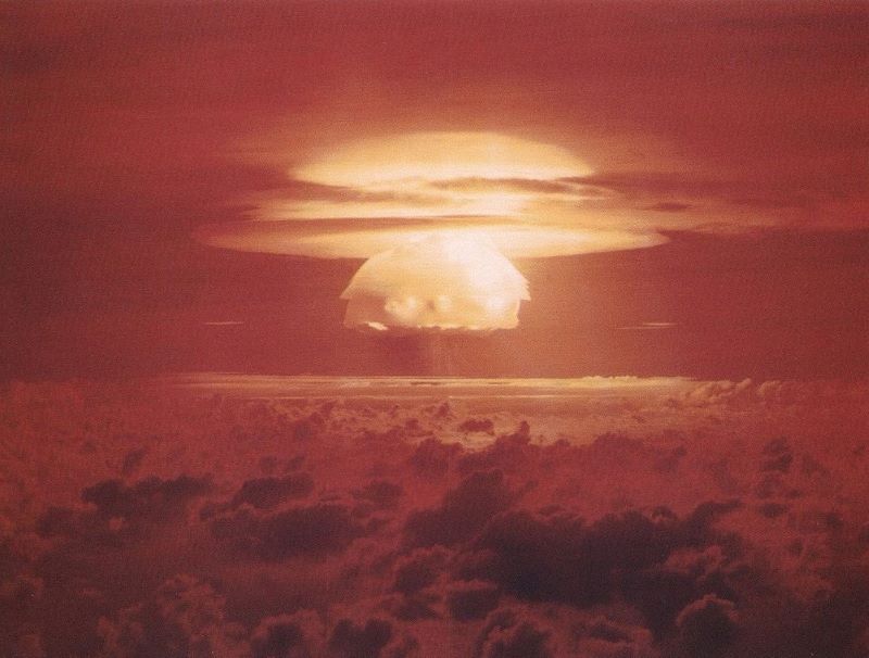 Nuclear war: Stubby white mushroom cloud seen between high and low clouds over ocean with red haze.