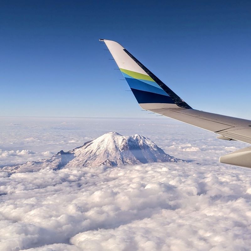 Airplane wing over snow-capped mountains with clouds below a conical mountain peak.