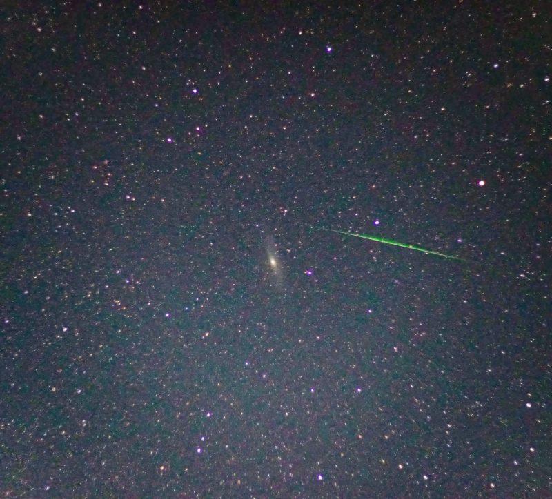 Small, yellowish, oval-shaped cloud with brighter center in star field with a nearby thin, bright green streak.