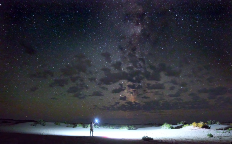 Man standing with light illuminating ground, with Milky Way and clouds above.