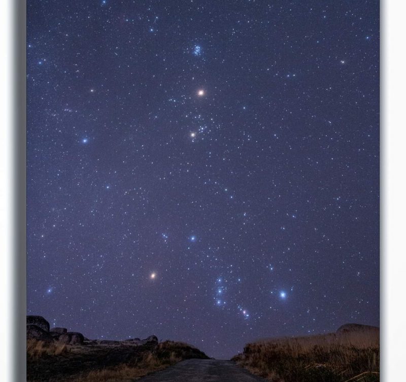 Starry sky dotted with brighter stars: Orion, Taurus, Pleiades, over rocky horizon, with red Mars.