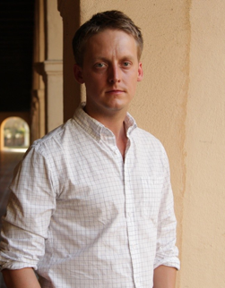 Serious young man standing with white button-down shirt, short brown hair.