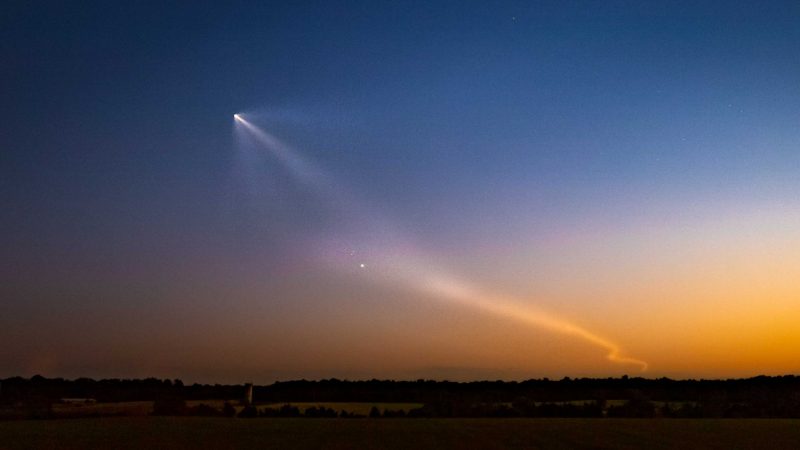 Colorful sunset sky with comet-like rocket exhaust leading down over horizon, and one bright planet.