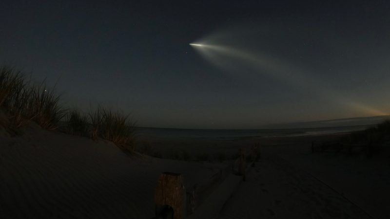 Cigar-shaped light of Starlink with billowing wispy light behind over dark beach and ocean.
