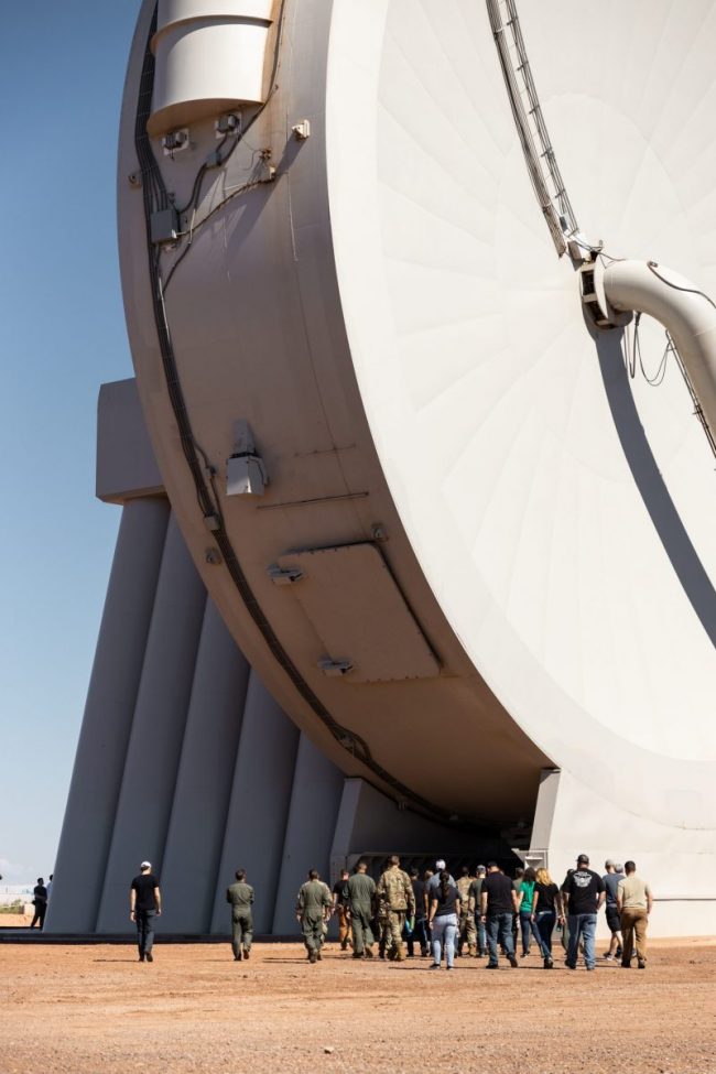 Group of people under large vertical disk shaped mechanical building.