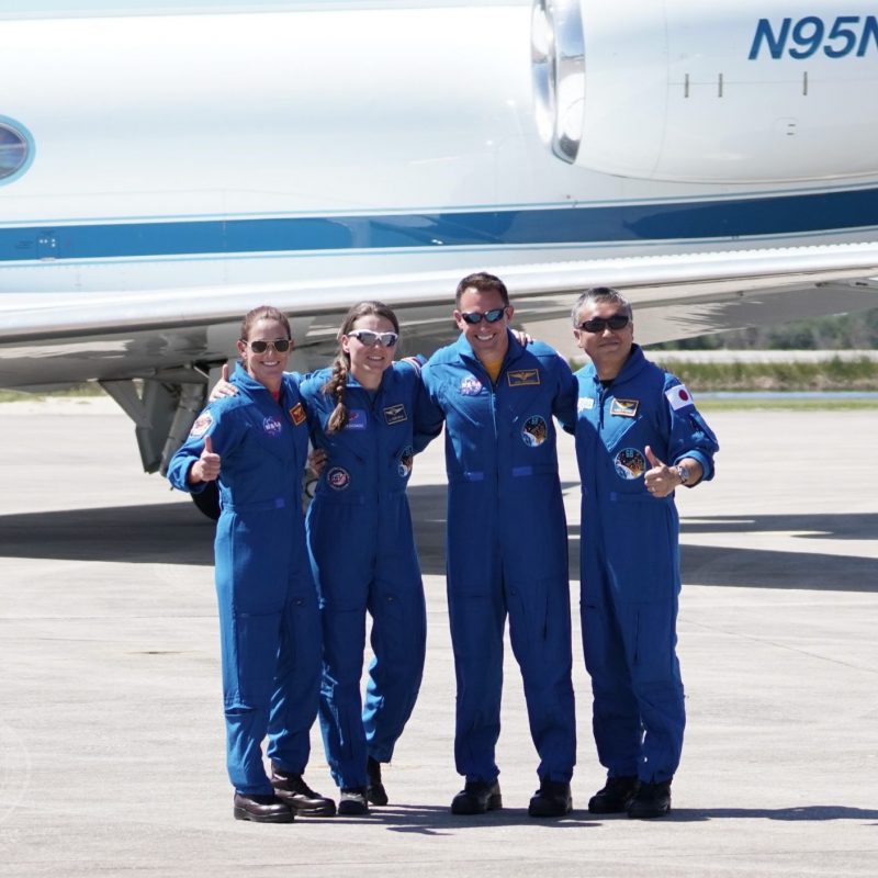 Two women and 2 men in blue jump suits, arms over each other's shoulders and thumbs up sign.