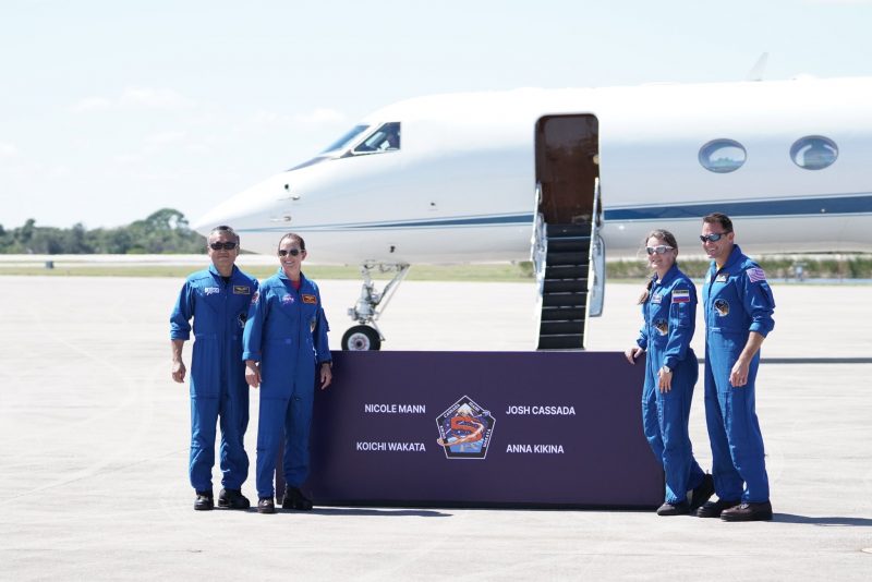 Four astronauts in blue jumpsuits, two on either side of a sign with their names and enlarged patch.