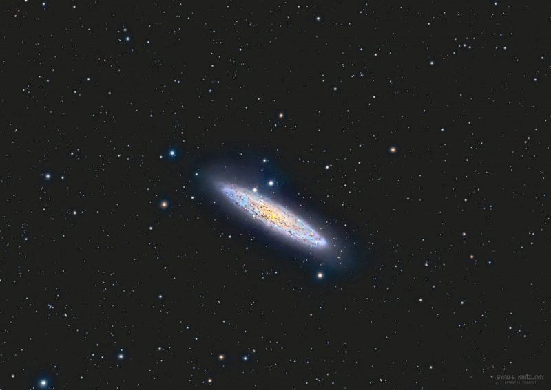 Very oblique view of medium-sized bluish spiral nebula with a yellow nucleus and foreground stars.