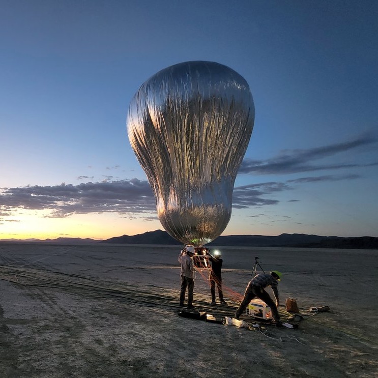 Venus balloon prototype: Large, oblong silver balloon being held by 2 people in a desert at sunrise.