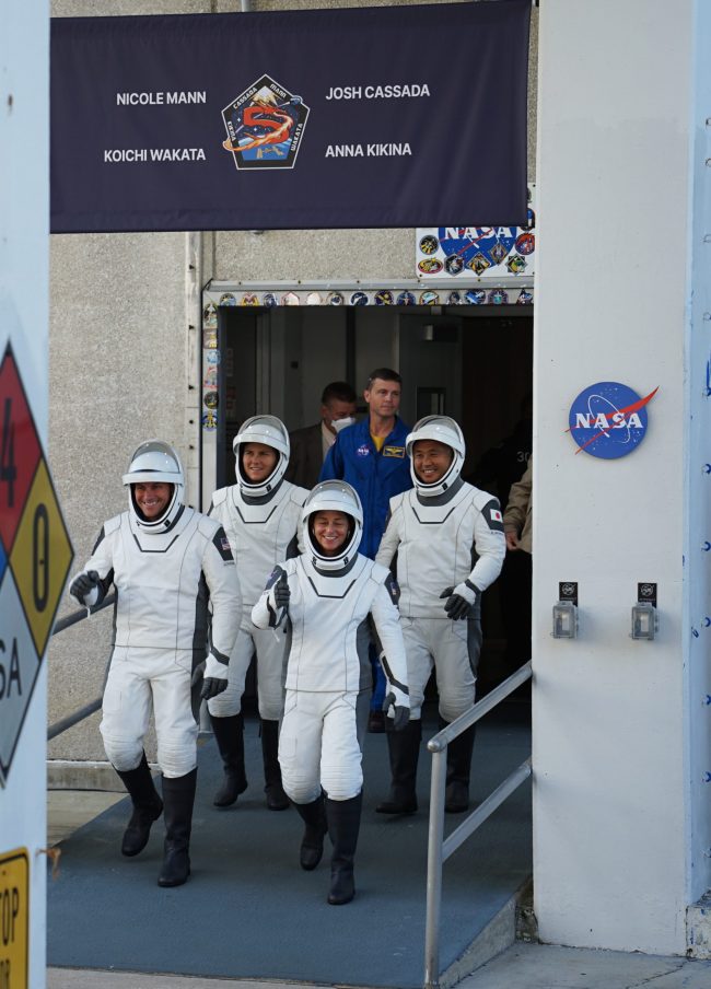 Launches: 4 people in spacesuits walk down a ramp outside while smiling.