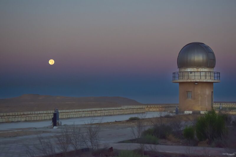 An observatory in a desert with the full moon in a pink band and dark blue below near the horizon.