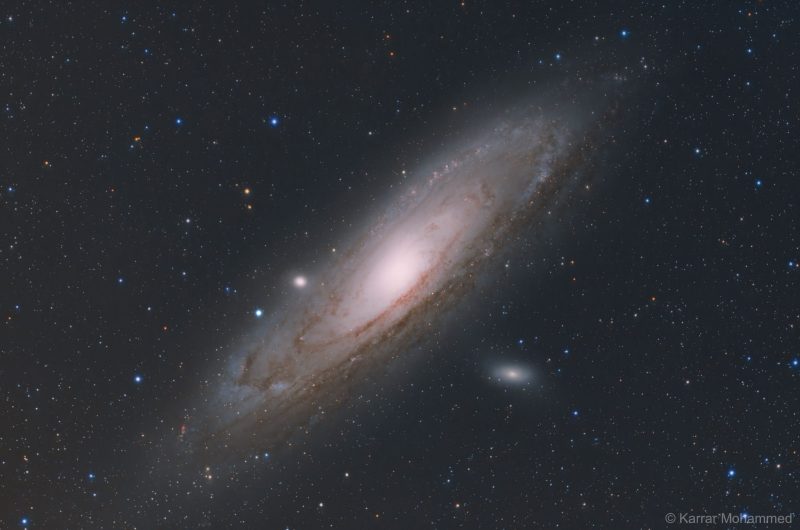 Oblique view of large yellowish spiral galaxy with 2 small glowing ovals and foreground stars.