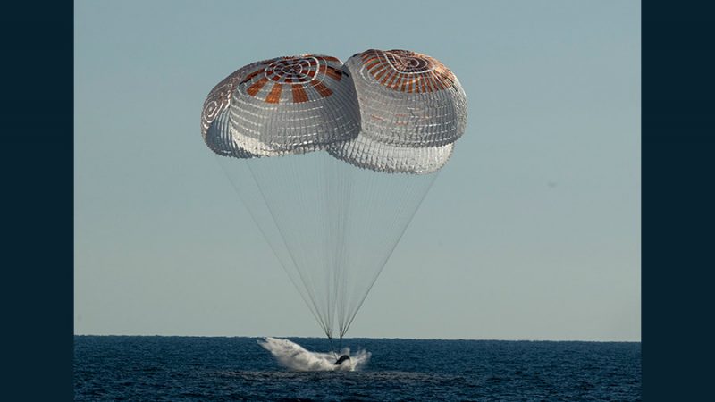 Two white parachutes above a space capsule, splashing down into slate-dark ocean.