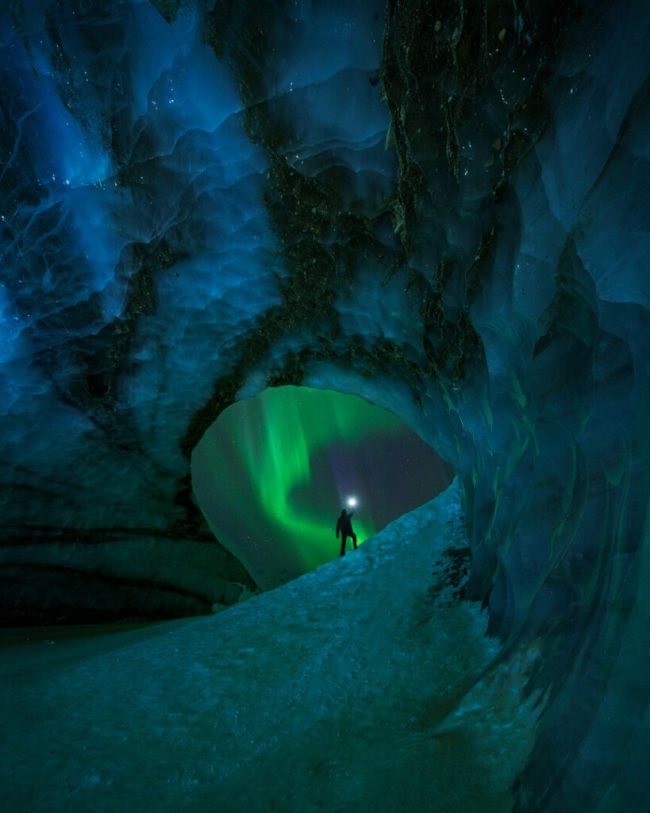 Man holds up a light inside an ice cave as green aurora is visible outside cave mouth.