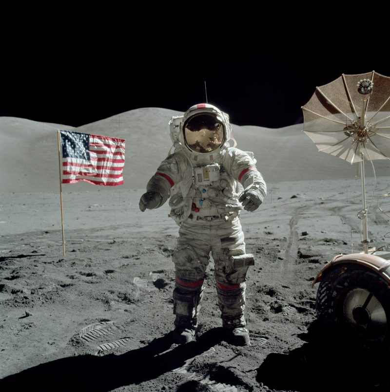 Apollo 17: Astronaut in spacesuit with gold helmet standing next to dish and American flag.